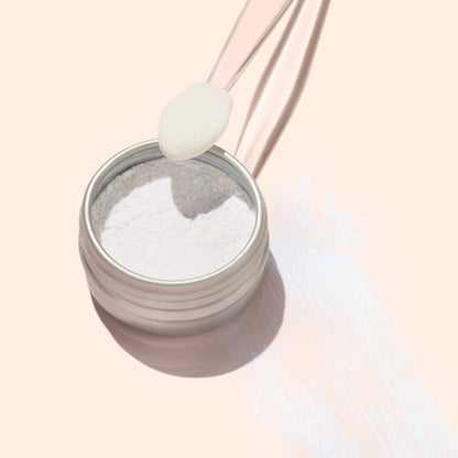 A small round tin of Manucurist Blue Glazed Powder rests on a peach surface. A translucent spatula holds a scoop above the tin, with pearly blue shimmer powder—ideal for adding brilliance to any manicure—scattered nearby.