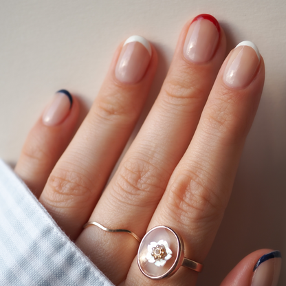 Une main aux ongles peints avec le Red, White Blue Trio de Manucurist — rouge, bleu marine et blanc — fait écho à l’audace d’un fard à paupières patriotique. Deux bagues dorées ornent les doigts : l’une fine, l’autre ornée d’un motif floral rond.