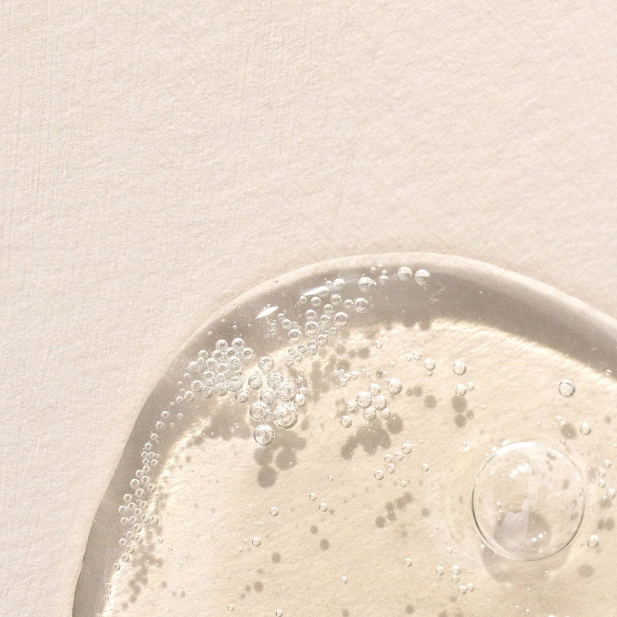A close-up of a translucent gel-like substance with bubbles on a light, textured surface. The gel appears glossy and is partially shown in the lower right corner of the image.