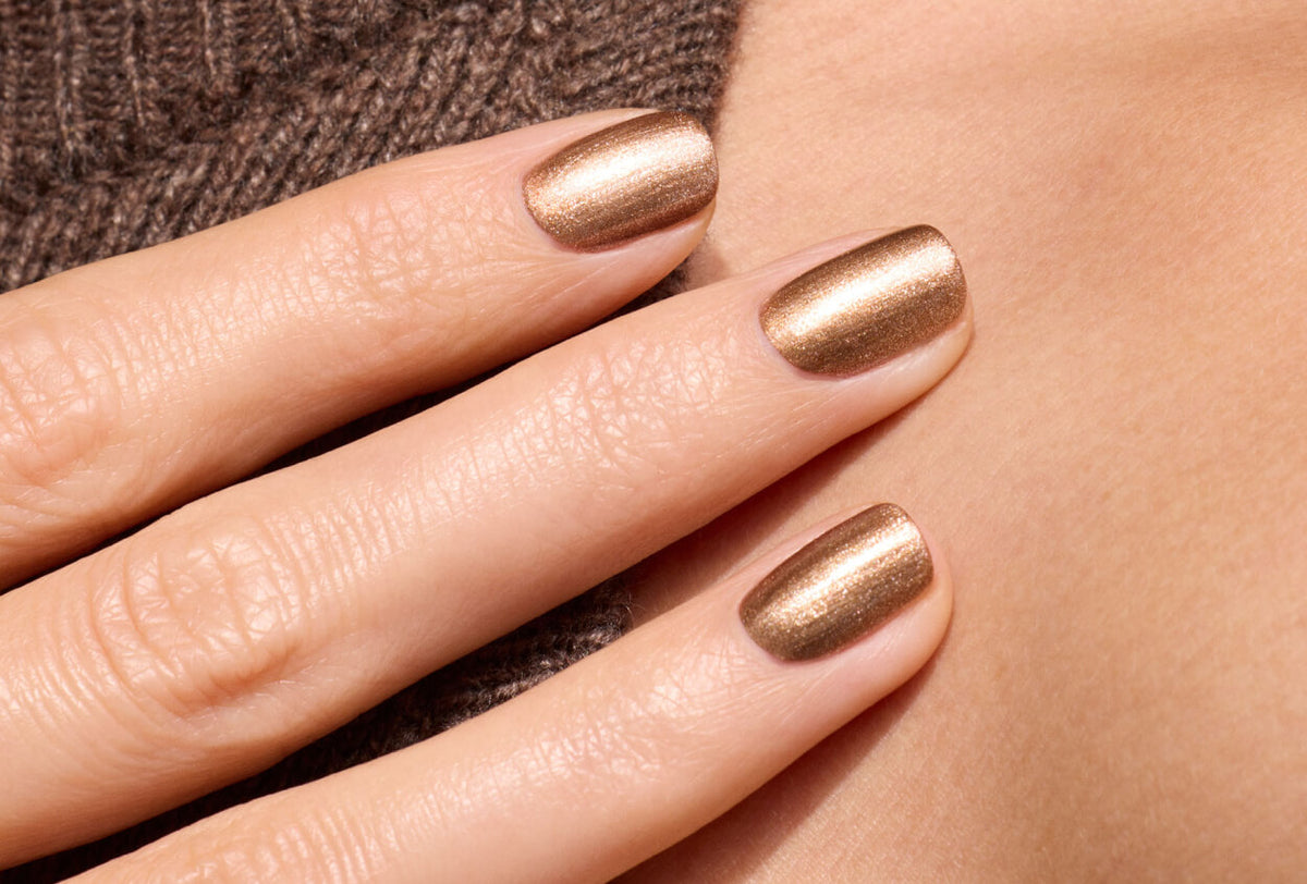 Gros plan d'une main avec un vernis bronze métallique, posée sur une peau nue et un tissu texturé marron. Les ongles sont parfaitement limés et le vernis offre un fini brillant et homogène.