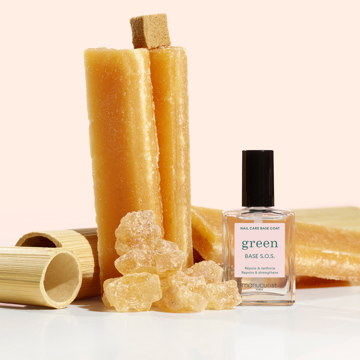 A bottle of Manucurist Green Base S.O.S. nail care sits in front of amber-colored sugar sticks, crystals, and bamboo tubes against a white background.