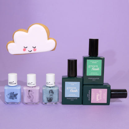 Cinq flacons du Set Manucurist You Me Under the Sea Green Flash™ sont disposés sur une surface violette. Trois teintes pastel aux étiquettes illustrées d’animaux sont mises en avant, avec un biscuit souriant en forme de nuage à l’arrière-plan.