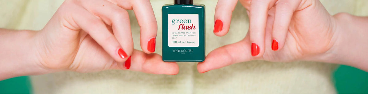 Close-up of hands with red-painted nails holding a green bottle of Manucurist Green Flash LED gel nail lacquer against a light background.