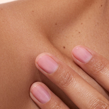 Here is the rewritten sentence using your product data: A close-up of three manicured fingers with pale pink nails, enhanced with Manucurist’s Active™ Glow Blueberry plant-based nail care, resting gently on a bare shoulder with smooth, light brown skin.