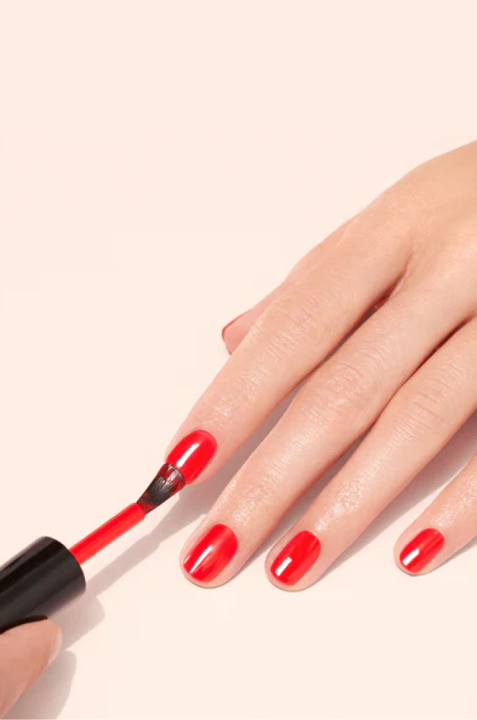 A hand is having its fingernails painted with bright red nail polish using a brush on a light background.