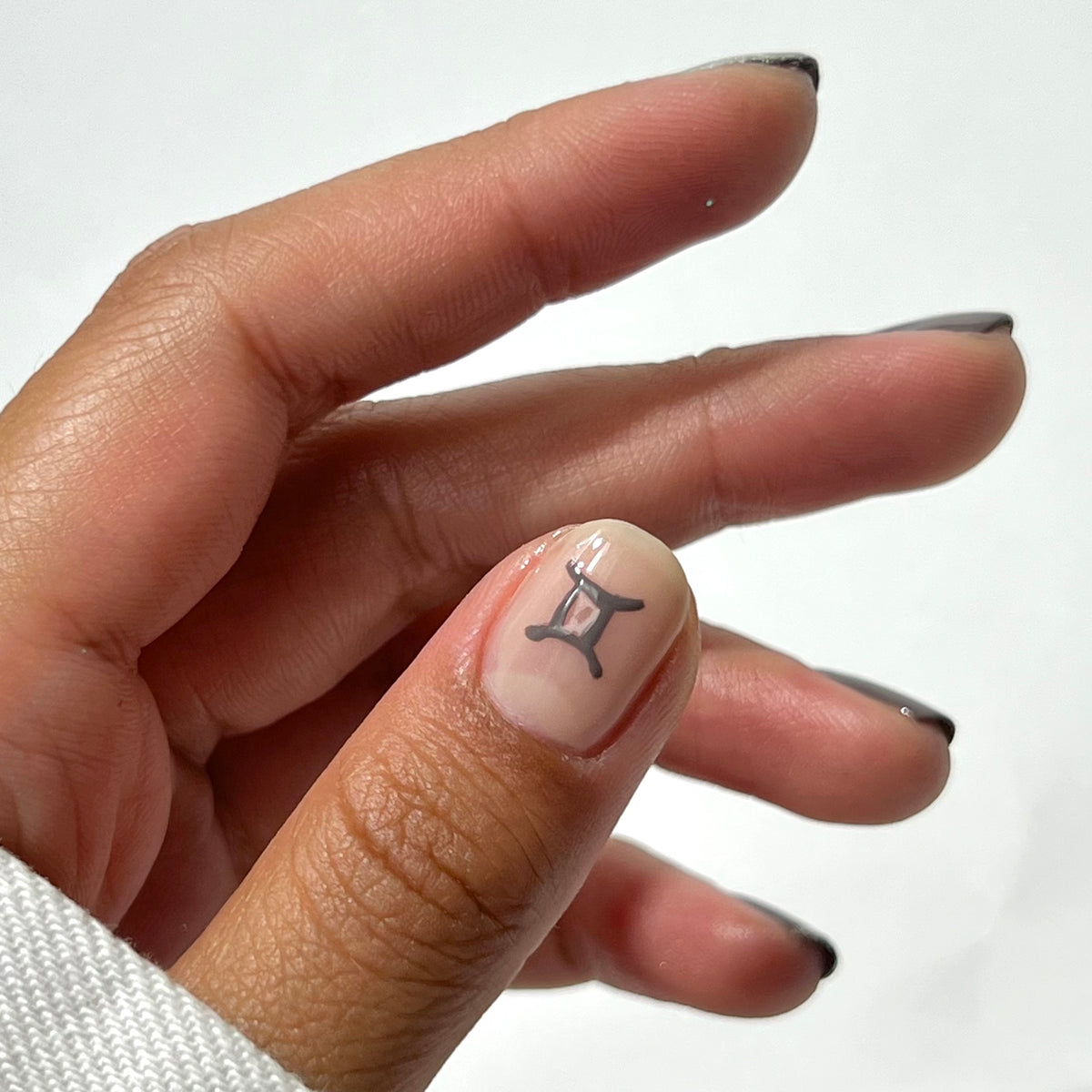 Une main aux ongles courts et soigneusement manucurés, avec un symbole astrologique des Gémeaux noir sur l’ongle du pouce. Les ongles ont une base transparente avec des pointes noires, et l’arrière-plan est blanc uni.