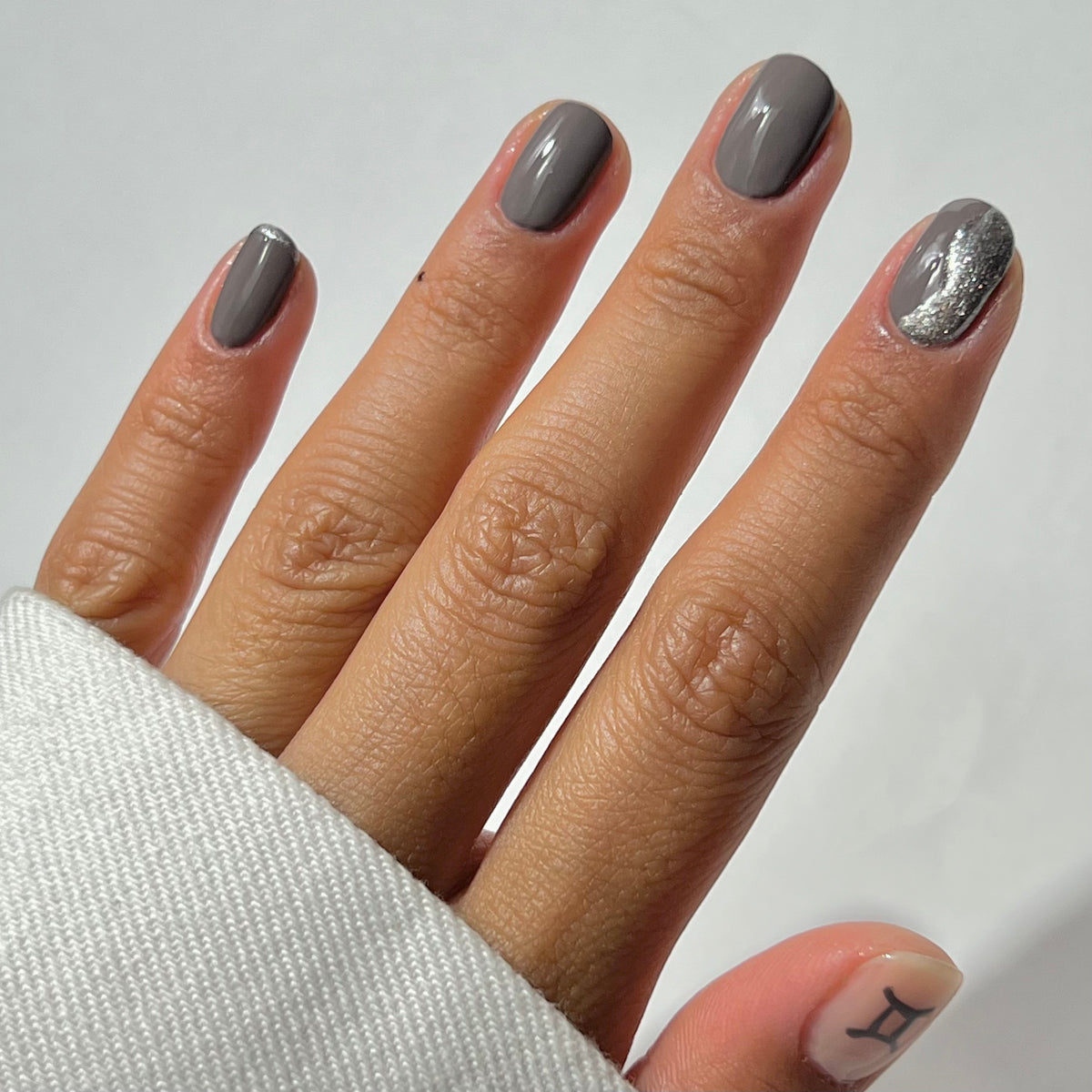 Une main aux ongles courts et soigneusement manucurés vernis en gris ; l'annulaire présente une touche pailletée argentée. Le pouce arbore un petit symbole du signe astrologique des Gémeaux. La personne porte un haut blanc à manches longues.