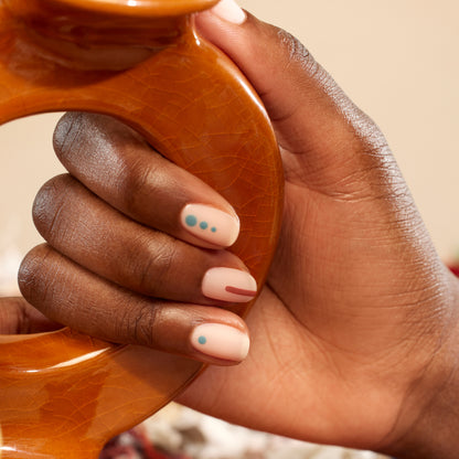 A hand with a light pink manicure from Manucurist’s Geometric Obsession features teal dots and a red line nail art while holding a brown, glossy circle.