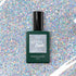 A bottle of Manucurist Mermaid Green Flash gel polish rests on a sparkling holographic glitter background that matches its glittery label.