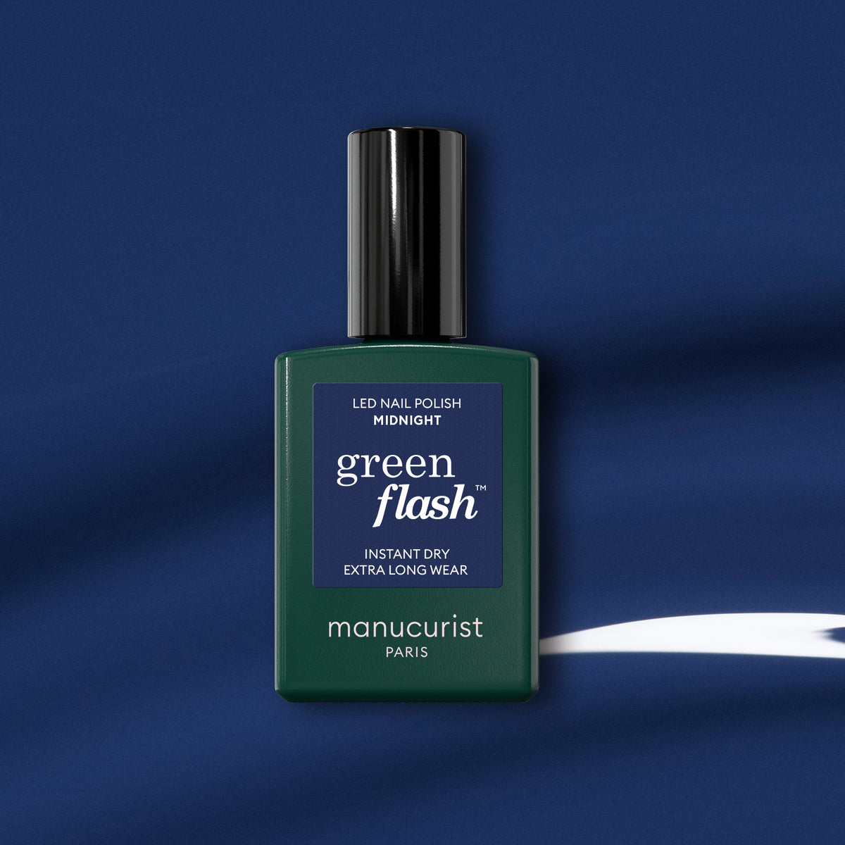 Un flacon de vernis gel Green Flash de Manucurist dans la teinte Midnight repose sur un fond bleu foncé avec une courbe blanche, avec un flacon vert foncé, un bouchon noir et une étiquette bleu marine mettant en valeur le vernis bleu nuit.