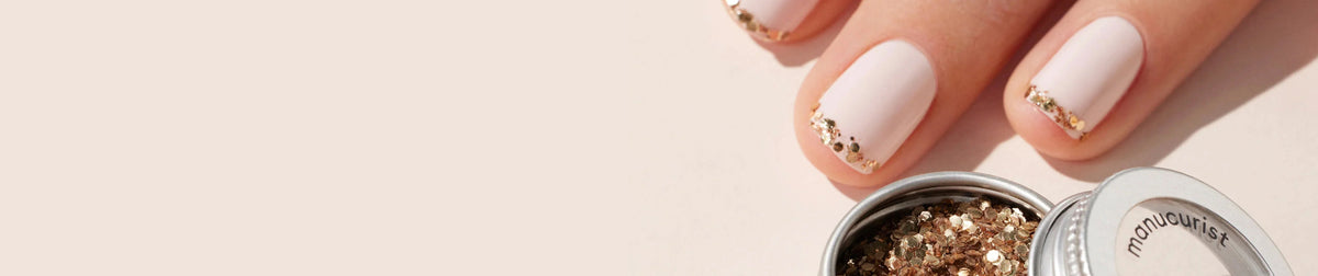 Close-up of a hand with light pink nails featuring gold glitter accents on the tips, next to an open container of gold glitter on a neutral background.