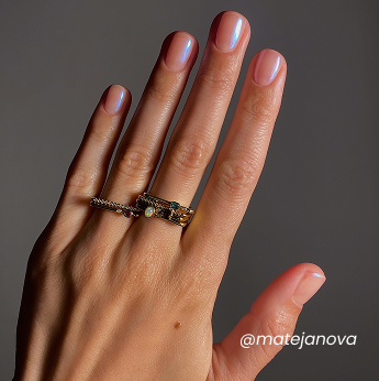 A hand with neatly manicured nails, painted with a light pink polish, wearing multiple gold rings with small gemstones. The background is plain and gray. The text matejanova appears in the lower right corner.