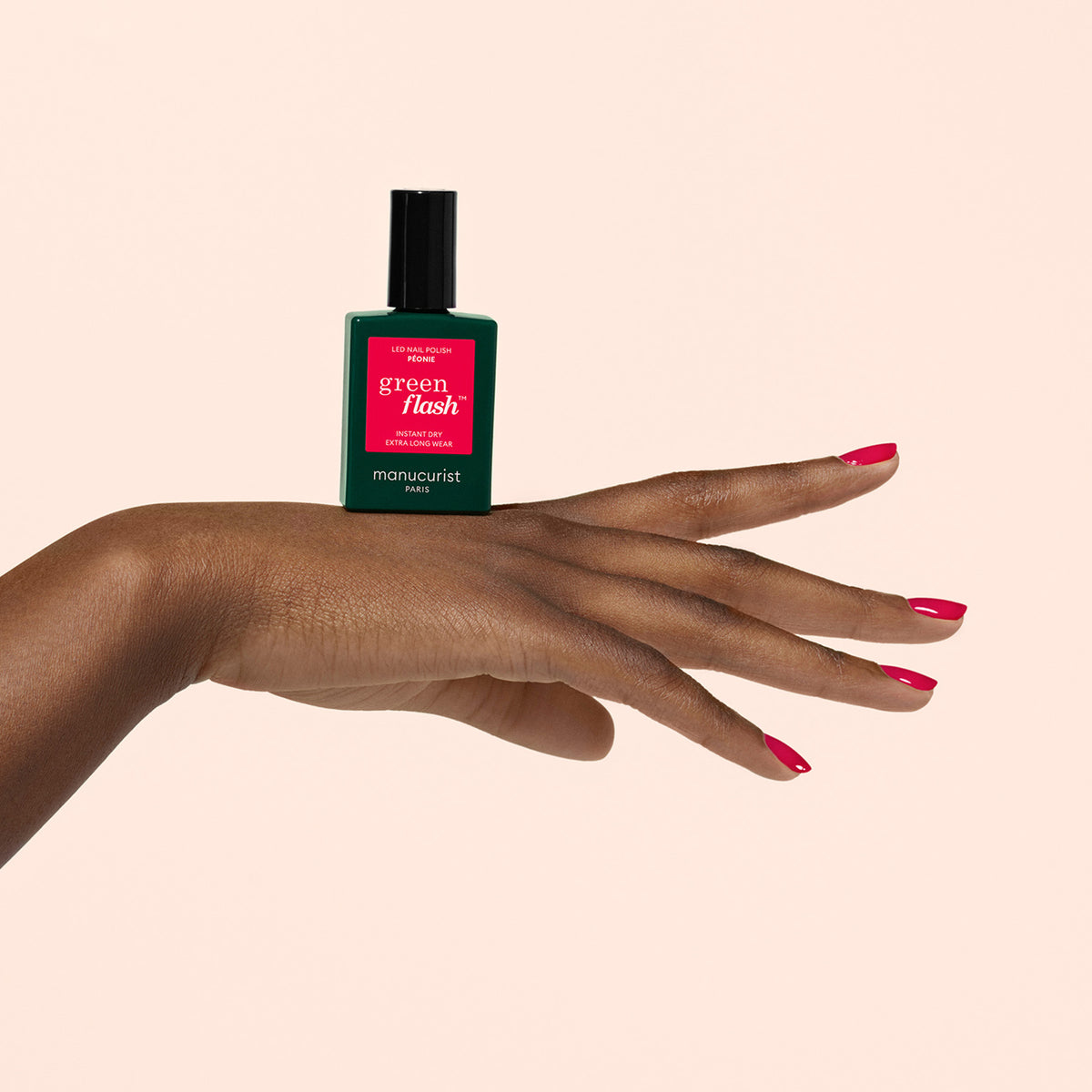 Une main aux ongles rouges vernis tient un flacon vert de vernis à ongles Manucurist Green Flash, en équilibre sur le dos de la main sur un fond beige uni.