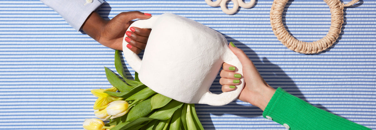 Two hands with painted nails hold a white vase together over a blue-and-white striped surface. Yellow tulips spill out of the vase, and rope decor items are visible nearby.