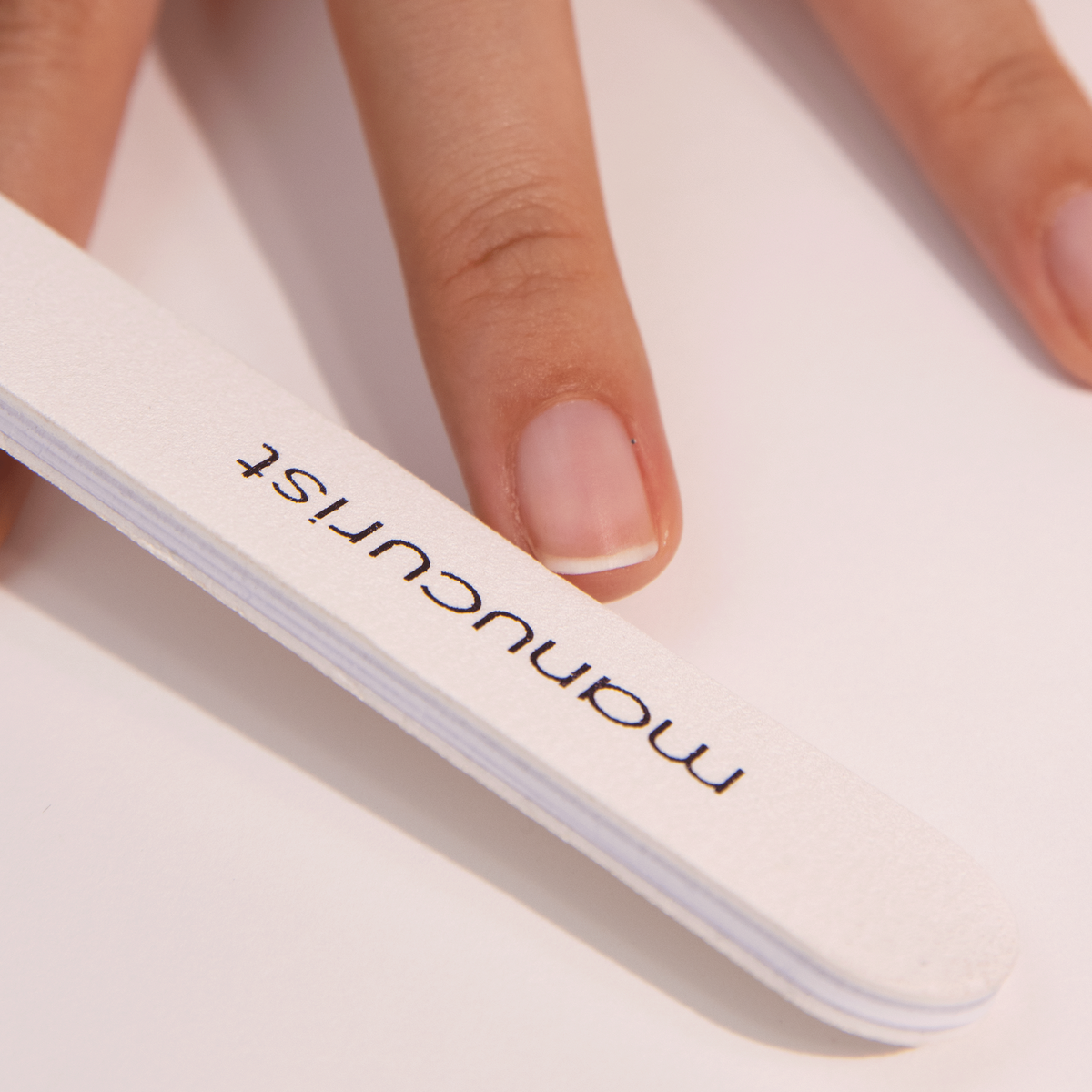 A close-up of a hand with natural nails holding a white nail file labeled manicurist on a light surface.