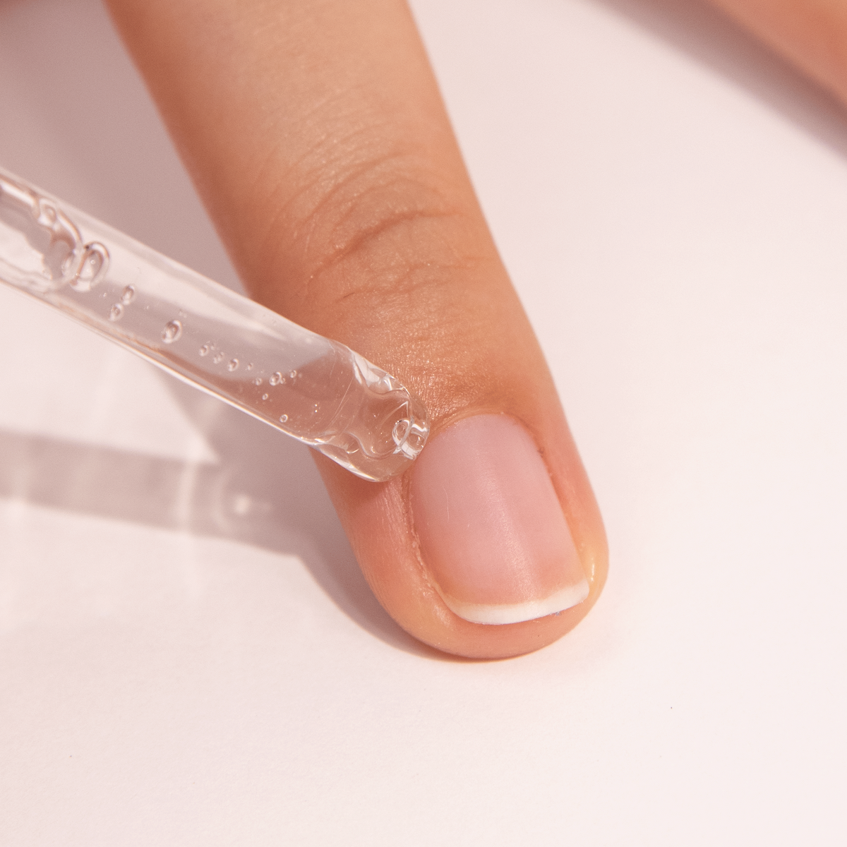 A close-up of a persons finger with clear liquid being applied to the nail cuticle using a dropper. The background is light and neutral.