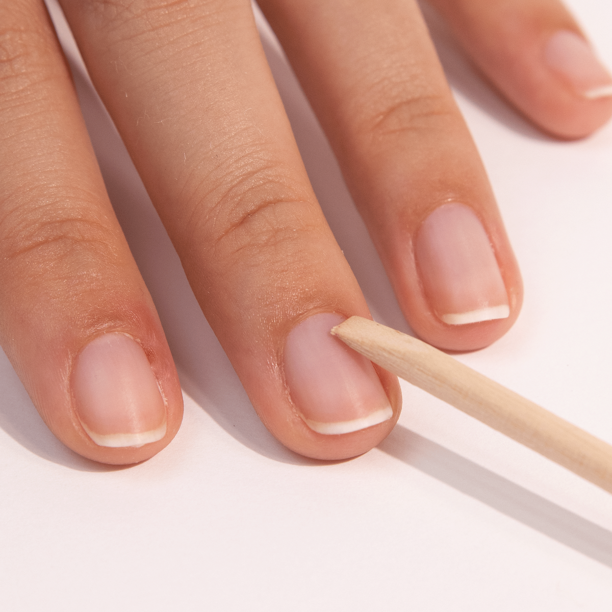 A close-up of a hand using a wooden cuticle stick to gently push back the cuticle on a fingernail, with natural, unpolished nails resting on a white surface.