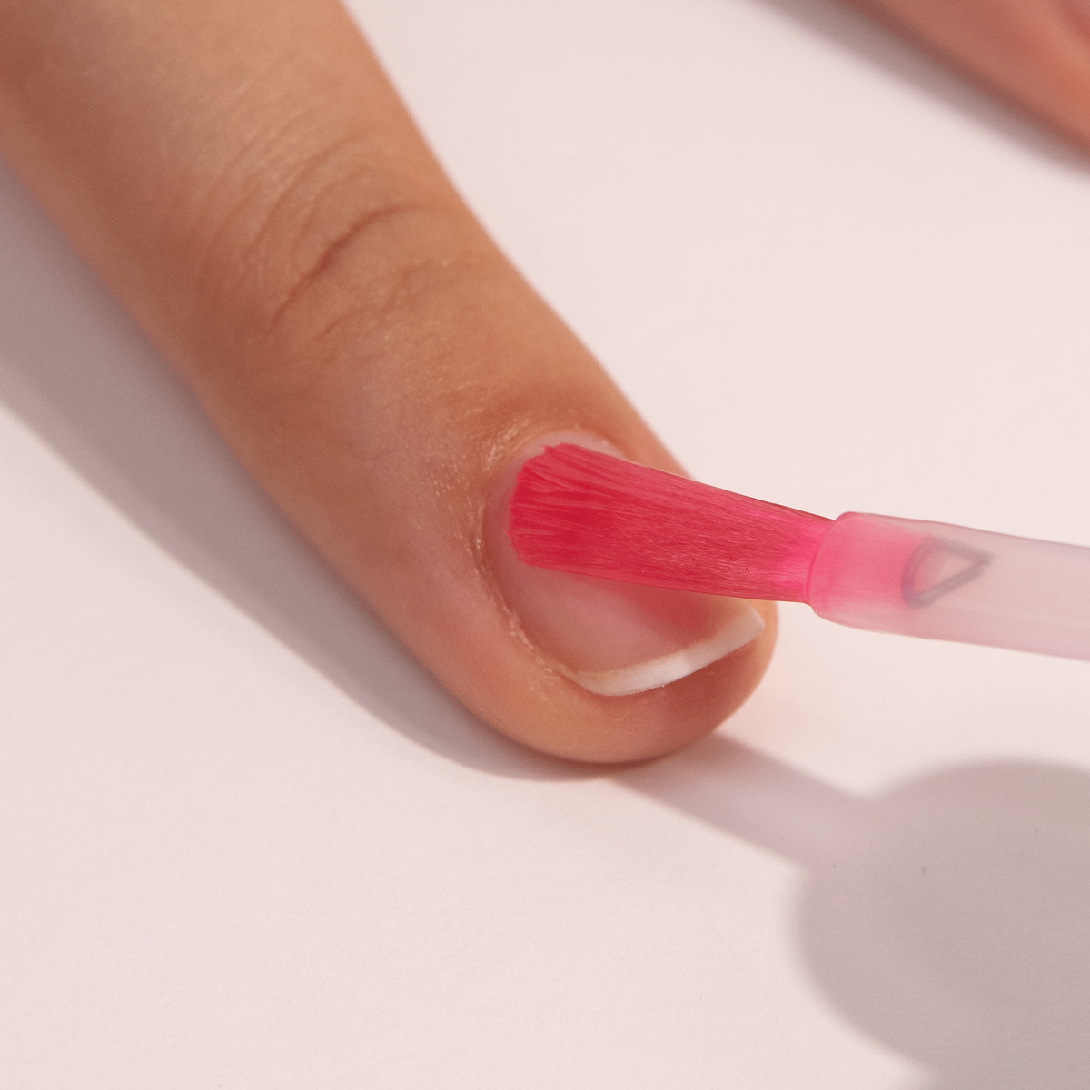 A close-up of a person applying pink nail polish to their fingernail with a brush, focusing on the nail and part of the finger against a light background.