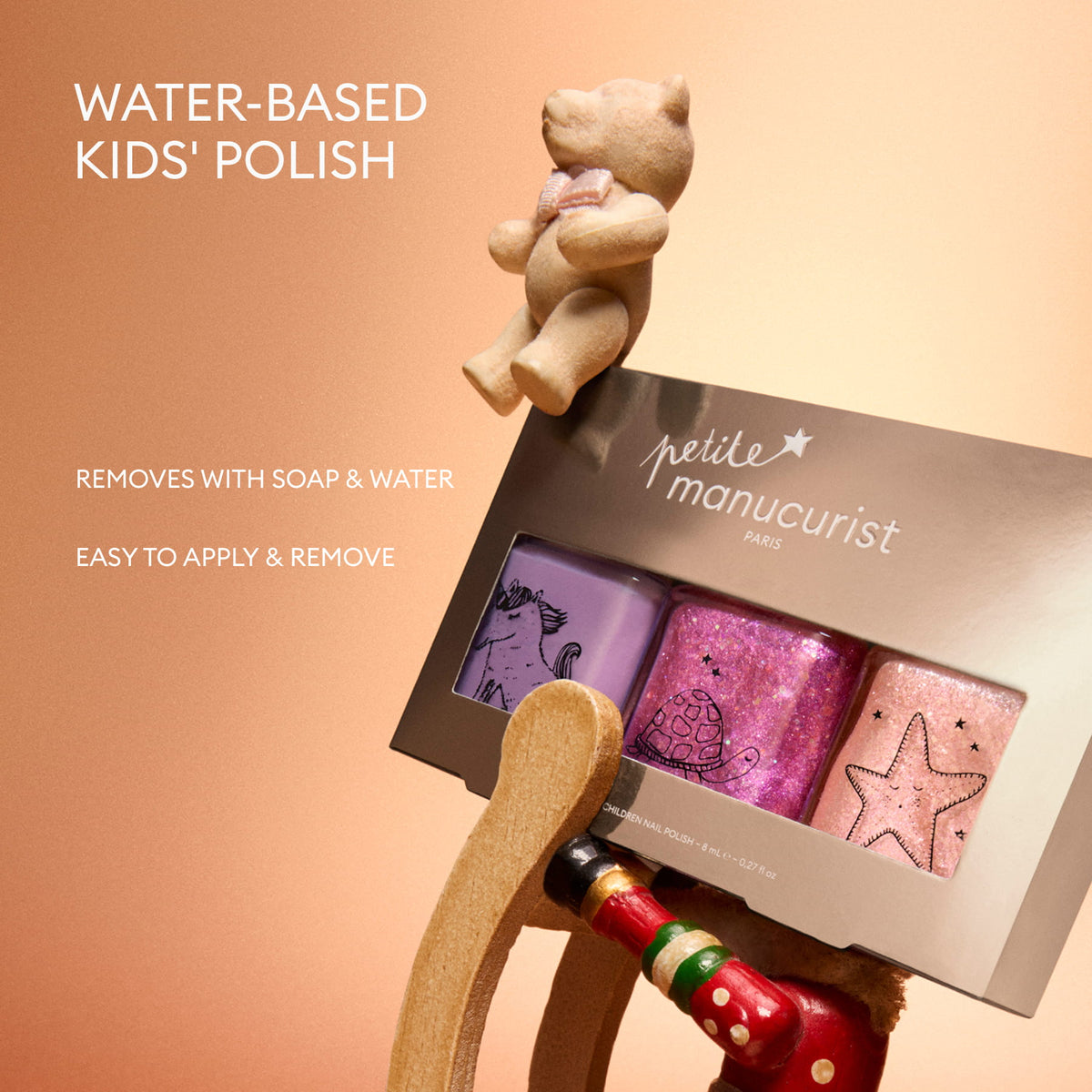 The Petite Manucurist Trio by Manucurist—a kids’ nail polish set featuring purple, pink, and glitter shades—is displayed on a rocking horse. Text reads: Water-based children’s nail polish of natural origin. Removes with soap  water. Easy to apply and remove—perfect as a holiday gift. A teddy bear is seated above.