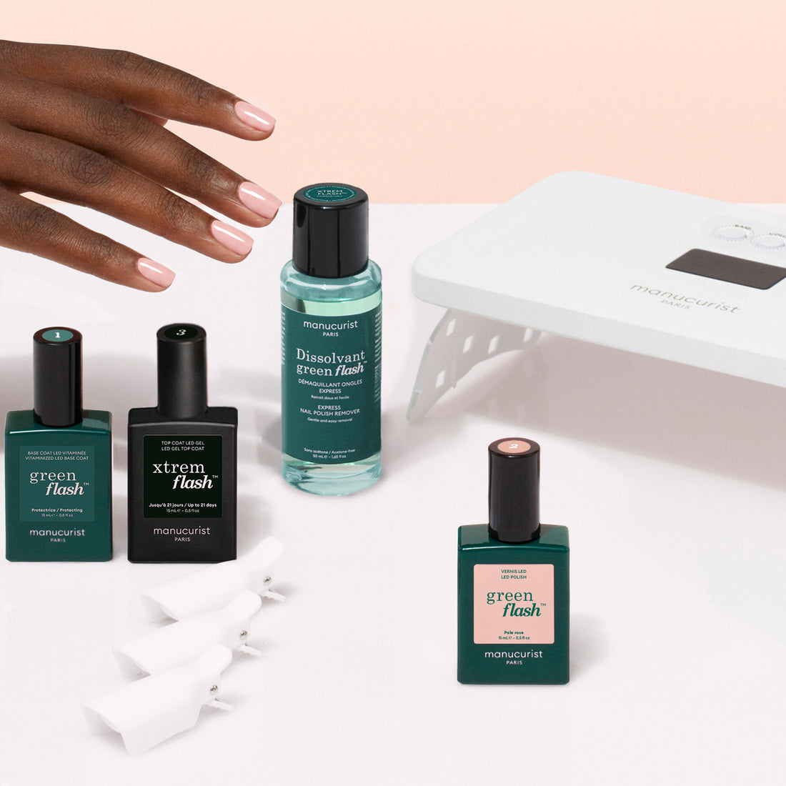 Une main aux ongles manucurés roses repose au-dessus d’un kit découverte Manucurist de vernis semi-permanent LED Soap Nails, comprenant trois flacons de vernis, du dissolvant, une lampe LED blanche et des pinces à ongles en plastique pour une manucure ultra-brillante.