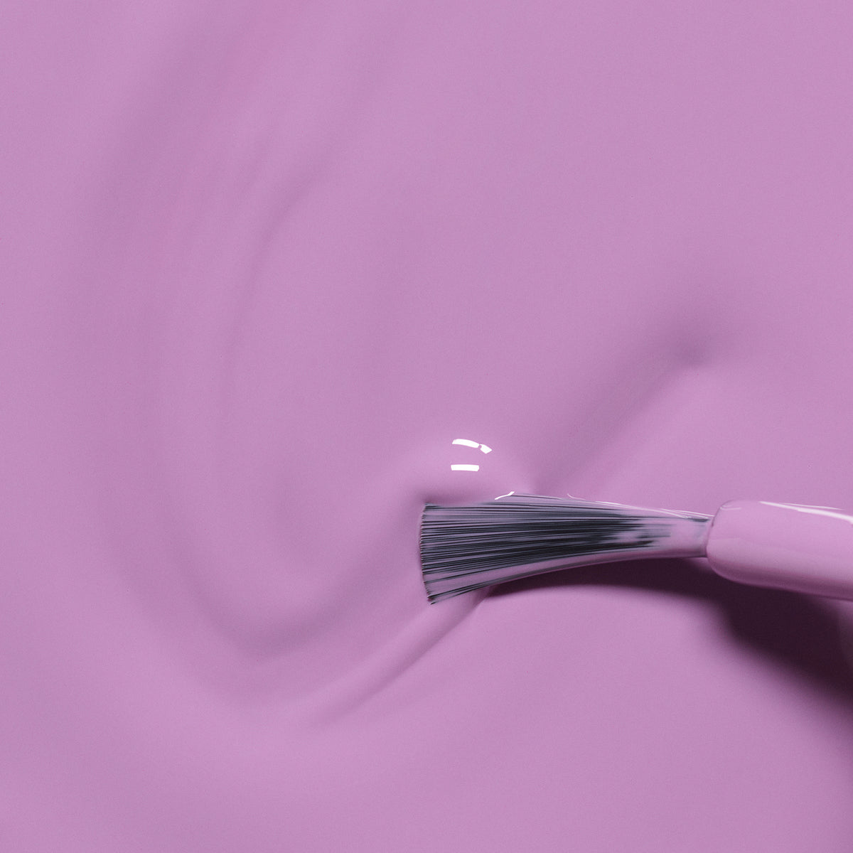 Gros plan du pinceau Manucurist Lily the Unicorn étalant un vernis violet lisse et brillant sur une surface, créant de douces volutes et des reflets.