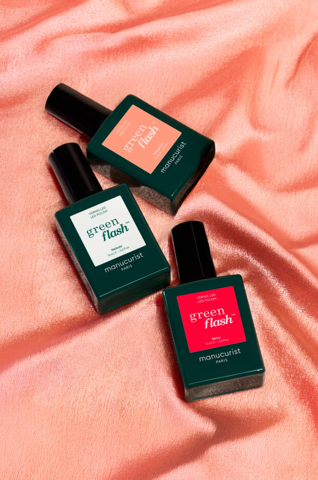 Three bottles of Manucurist Green Flash nail polish on a peach-colored satin fabric. The bottles are rectangular, dark green, with different colored labels: coral, white, and bright red.