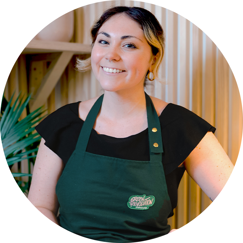 A smiling woman with short hair wears a black top and a green apron with a Green Revolution logo, standing in front of a light-colored, striped background with a plant to the side.