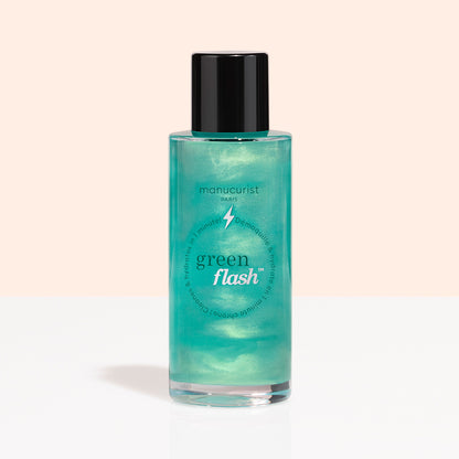 Un flacon transparent avec un bouchon noir, étiqueté « Manucurist Green Flash™ Gentle Glitter Remover », contient un liquide bleu sarcelle scintillant, sans acétone, et est présenté sur un fond pêche clair et blanc.