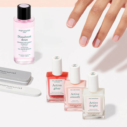 A manicured hand is displayed above bottles of nail treatments, nail polish remover, and manicure tools from the Active Manicure Kit by Manucurist, all neatly arranged on a white surface.