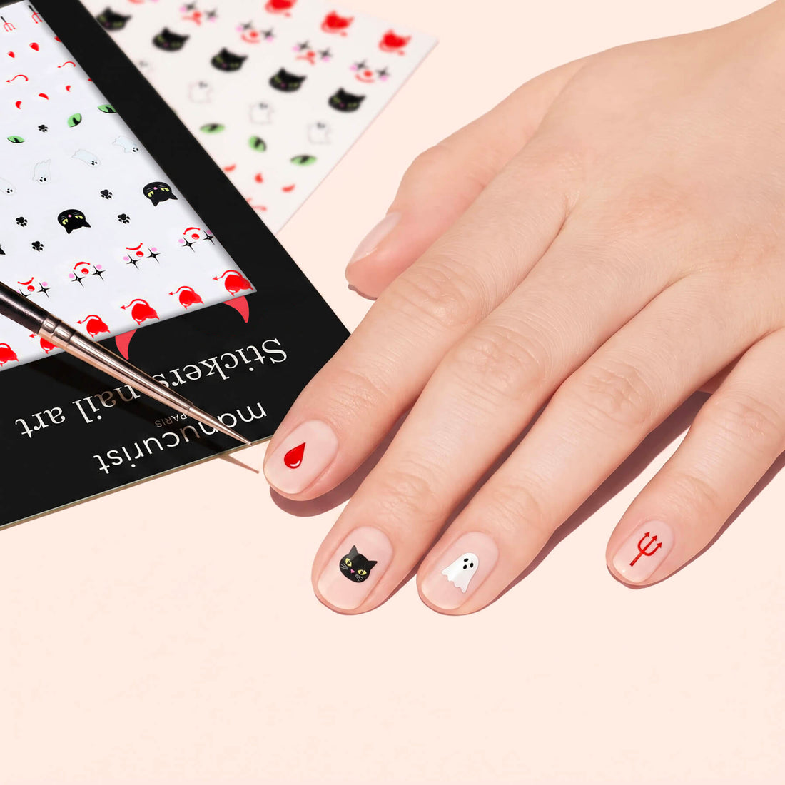A hand decorated with Manucurist Halloween Nail Art Stickers—red heart, black cat, ghost, and red trident—while a brush points to the nails and a sticker sheet with spooky designs is nearby.