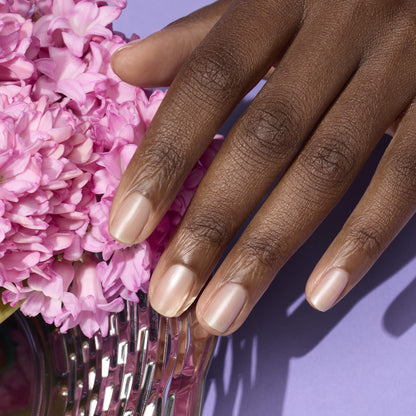 Une main manucurée avec soin, vernie avec Pearlescent de Manucurist dans une teinte nude naturelle aux sous-tons roses, posée à côté de fleurs roses sur une surface violette.