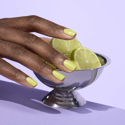 A hand with Manucurist Lime nail polish rests on a metal bowl filled with lime slices, set against a pastel purple background.