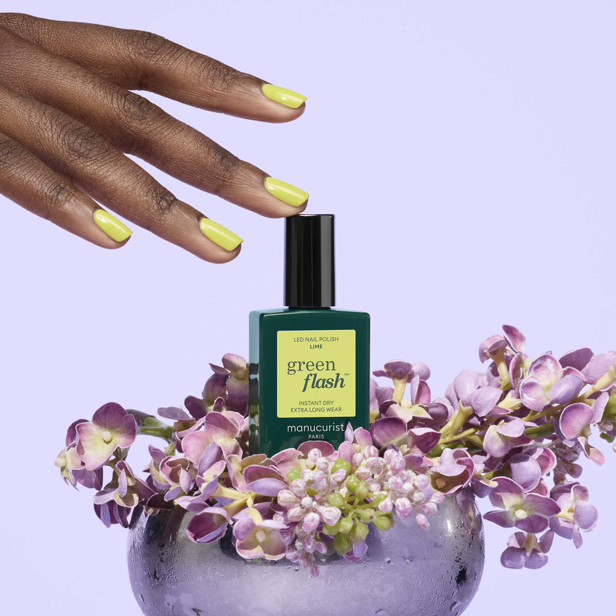 A hand with yellow-green painted nails hovers above a chartreuse Manucurist Lime gel polish bottle, surrounded by purple flowers in a clear vase, set against a pale lavender background.