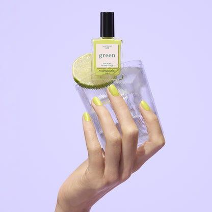A hand with chartreuse green Manucurist Lime polish holds a glass of water with a lime slice, topped with a matching Manucurist Lime bottle, all set against a light purple background.