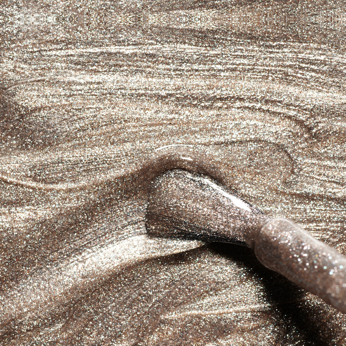 Gros plan sur le pinceau de maquillage Metallic de Manucurist tourbillonnant dans une poudre bronze scintillante, créant des traînées texturées et pailletées aux reflets métalliques froids.