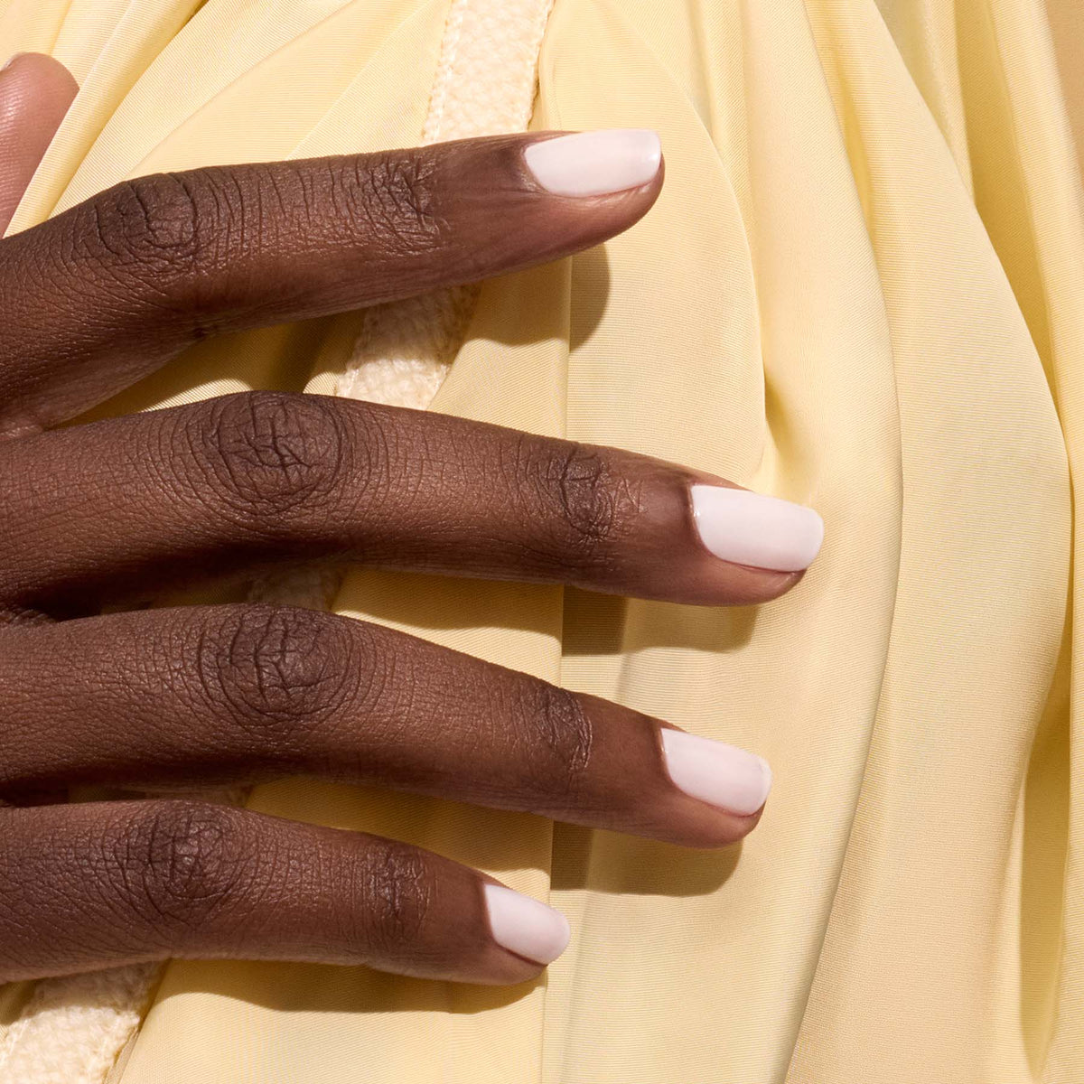 Gros plan d’une main à la peau moyennement foncée, aux ongles soigneusement manucurés et vernis en Milky White de Manucurist, posée sur un tissu doux jaune pâle.