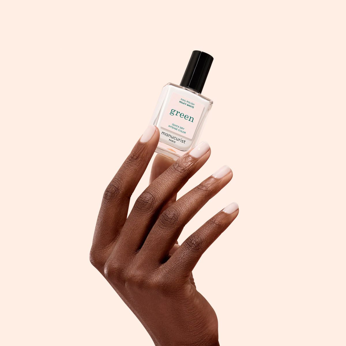 Une main aux ongles soigneusement manucurés tient un flacon de vernis à ongles Milky White de la gamme Green™ de Manucurist sur un fond beige clair.