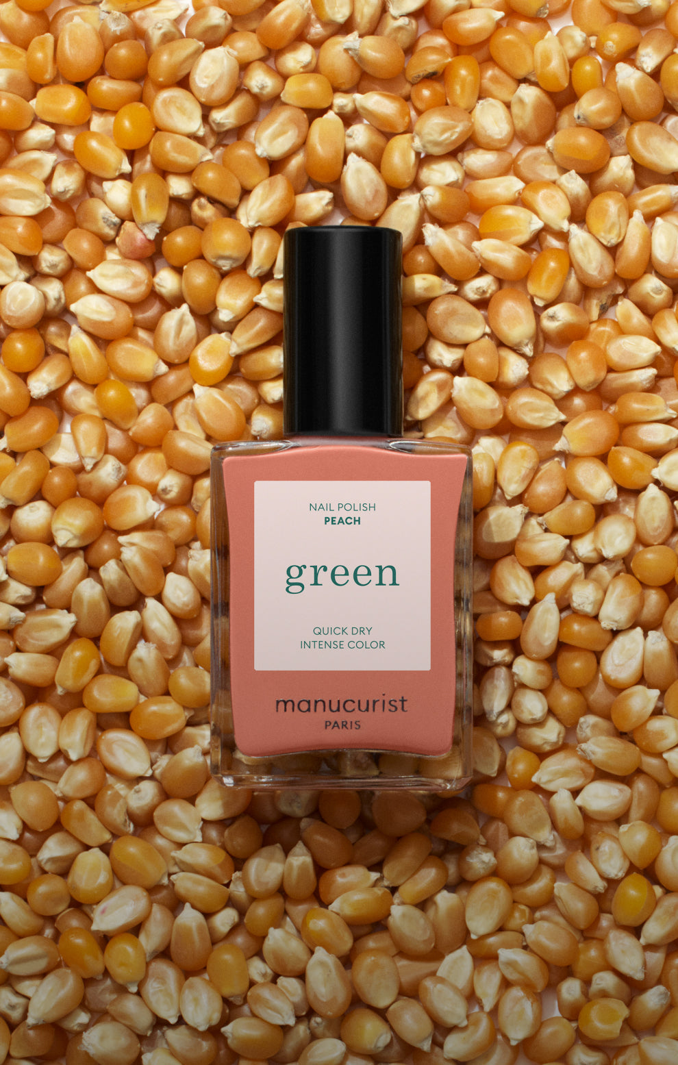 A bottle of peach-colored nail polish labeled green by Manucurist Paris rests on a bed of yellow popcorn kernels.