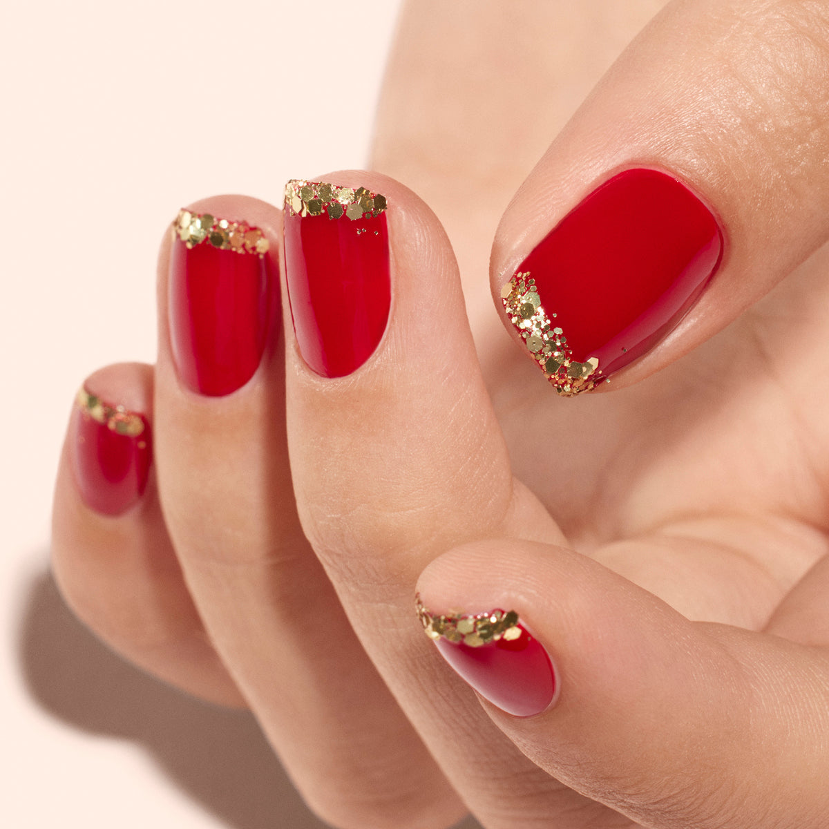 A close-up of short, rounded nails painted with Manucurist’s Ready to Party Duo, featuring Red Velvet polish and Gold biodegradable glitter tips—a festive, elegant manicure ideal for holiday gifting.