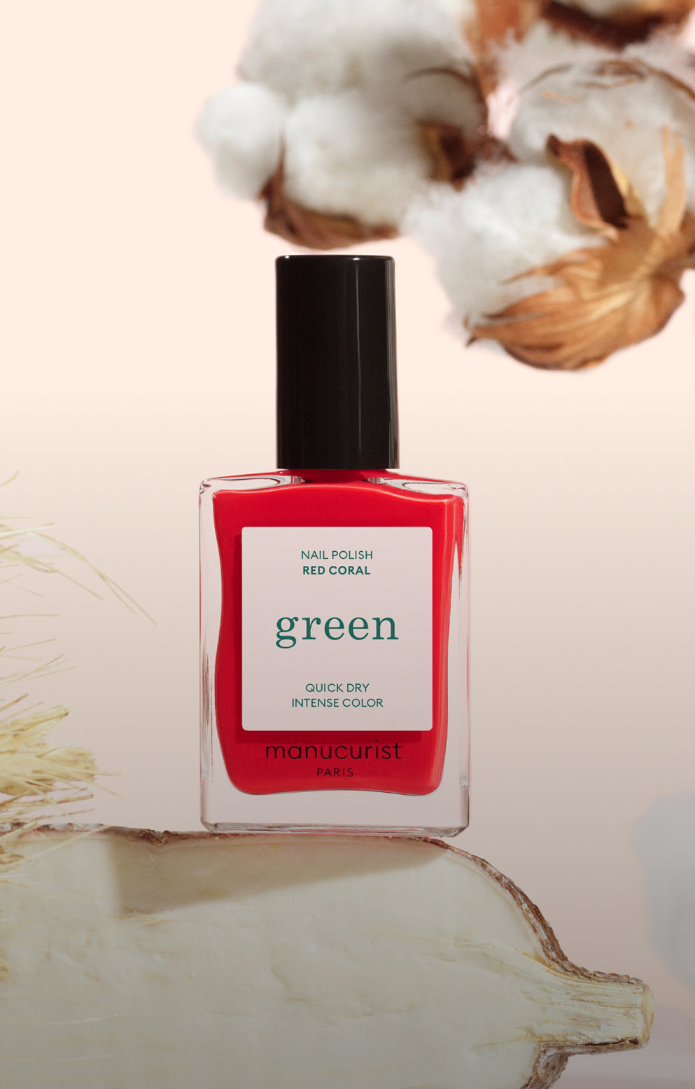 A bottle of Manucurist Paris Green nail polish in the shade Red Coral, with a black cap, is placed in front of a beige background with cotton and plant elements.