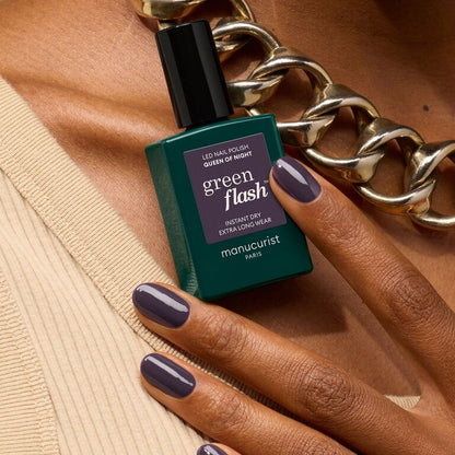 Une main aux ongles gris violacé foncé tient un flacon de vernis Manucurist Queen of Night, ornée d&