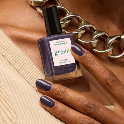 A person with dark skin, in a beige top and chunky gold chain, holds Manucurist’s "Queen of Night" nail polish. Their nails are painted the same rich deep purple as the shade in the bottle.