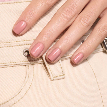 A close-up of a hand with short, neatly manicured nails painted in Manucurist’s Old Rose, a glossy antique pink polish, resting on the pocket of beige denim pants.