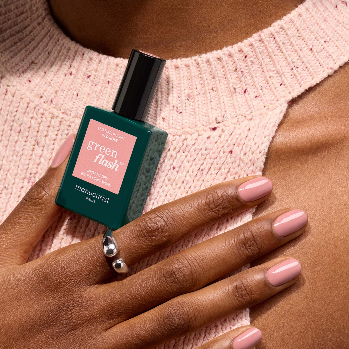 A hand with Old Rose gel polish by Manucurist rests on a textured pink sweater, holding the bottle. The person wears a silver ring with two spheres.
