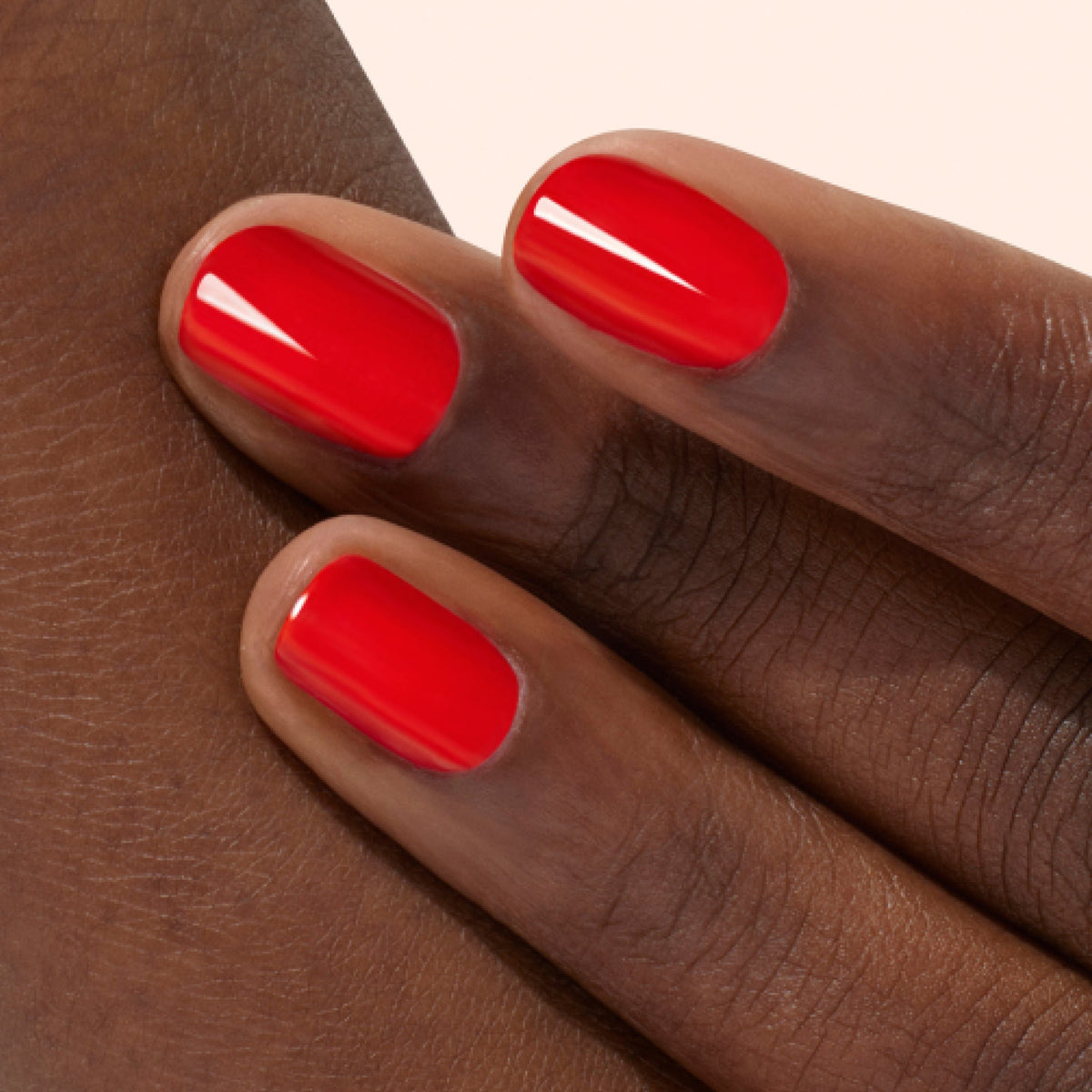 Close-up of three fingers with dark skin, featuring neatly manicured nails painted with glossy bright red nail polish. The fingers are gently resting on the back of a hand.