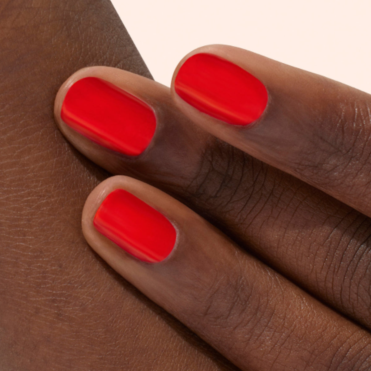 A close-up of three fingers with smooth, dark skin, featuring neatly manicured nails painted with glossy, bright red nail polish. The background is a soft, pale color.