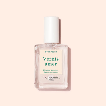A clear rectangular bottle of Manucurist Bitter Polish with a white cap rests on a pale pink background. Its cream label with green and beige text highlights nail care benefits, helping prevent nail biting.
