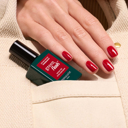 A hand with glossy Manucurist Pomegranate gel polish holds a bottle of Green Flash by Manucurist, partially tucked into the pocket of a beige textured garment.