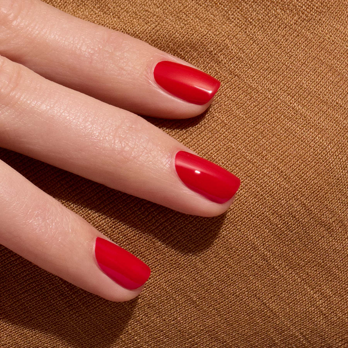 A close-up of a hand with neatly manicured nails painted in Manucurist’s glossy Poppy Red polish, resting on a textured brown fabric.