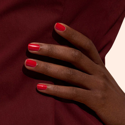 A hand with dark skin and neatly manicured nails painted in Manucurist’s Poppy Red rests against a burgundy fabric background.